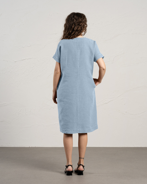 The back view of a woman with curly brown hair shows them wearing a cloudy blue linen dress with short sleeves and pockets, standing against a textured off-white wall.
