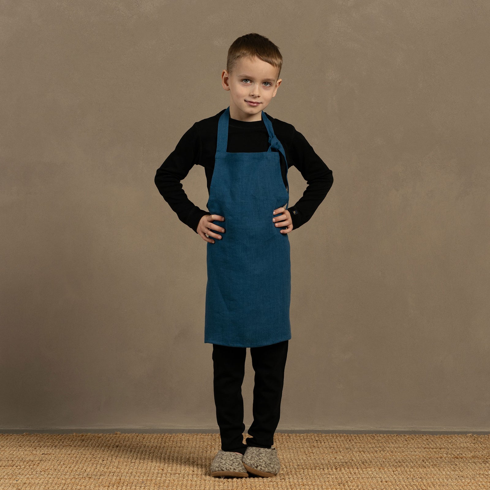 A child wearing a linen apron with leaf dots, standing at a table involved in crafting or baking, with ingredients and baking tools on the table.