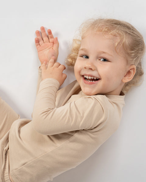 Laughing child lying on the floor in beige bamboo long-sleeve top and bottoms.