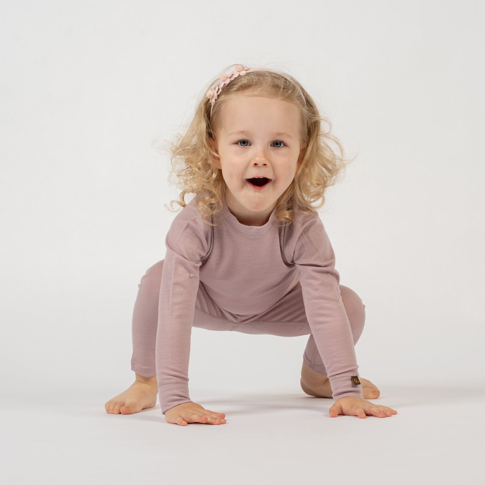 A child with curly blonde hair and a light pink floral headband stands against a white background, wearing a dusty pink long-sleeved top and matching pants. The child has one hand on their hip and is looking at the camera with a slight smile.