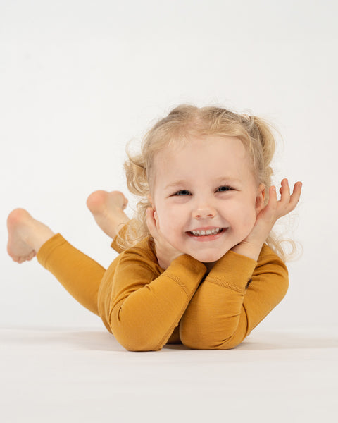 A happy child with curly blonde pigtails, wearing a yellow long-sleeved top and matching pants, is lying on their stomach on a white surface. The child is propped up on their elbows with their chin resting in their hands, smiling widely at the camera. Their bare feet are visible in the background.