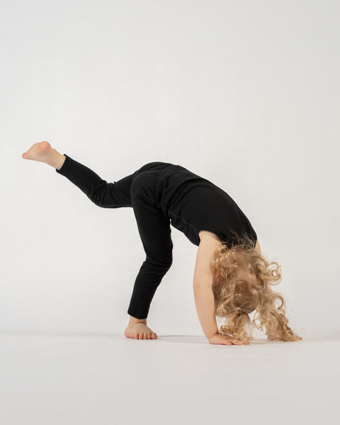 A child with blonde, curly hair, wearing a black sleeveless top and black pants, is performing a playful pose. They are bent over with their hands on the white floor, and one leg is lifted straight up behind them.