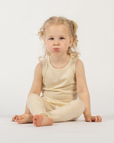 A young child with light curly hair in pigtails is sitting on a white surface against a plain white background. The child is wearing a menique sleeveless top and matching leggings. They are looking directly at the camera with a pouty expression, and their bare feet are visible.