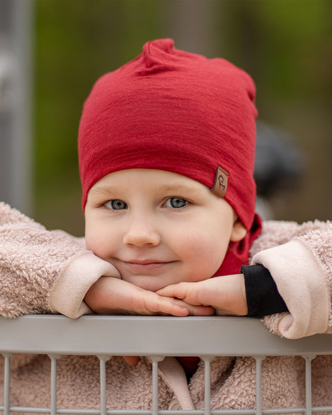 A young child with light skin and blue eyes is looking directly at the camera with a slight smile, resting their chin on their hands. They are wearing a menique royal cherry beanie and a light-colored, textured coat, leaning over a light gray railing or barrier.