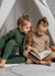 Two little girls sitting in an inside play tent, reading a book. One of them is wearing a merino wool base layer set in dark green, and the other one in creamy beige color.