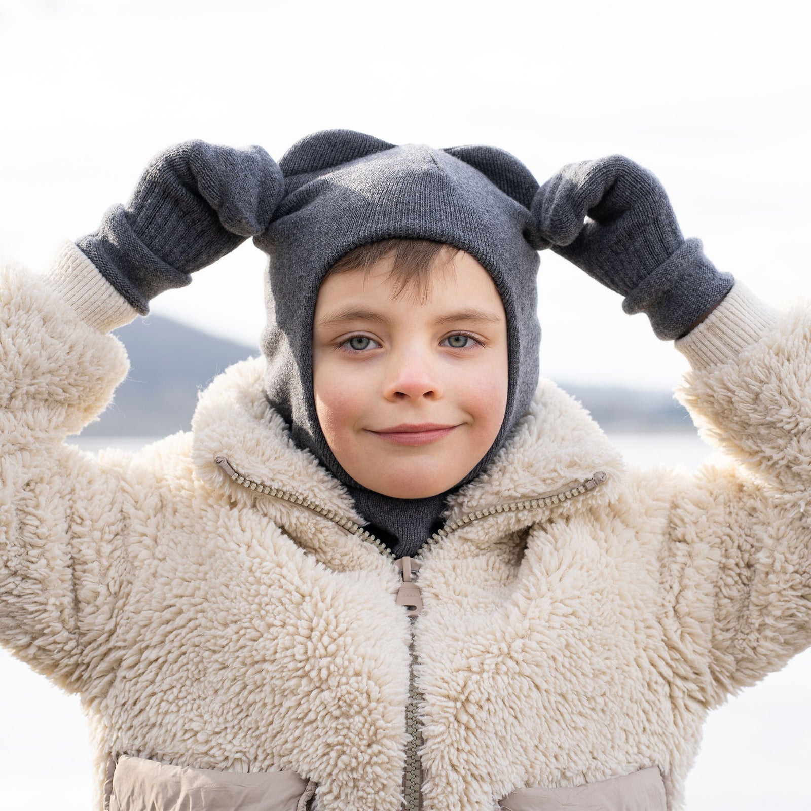 A child with light brown hair and blue eyes wears a dark gray knit balaclava with two small, rounded ear-like protrusions on top. They also wear a light-colored, fuzzy or shearling-like jacket with a zipper partially open, revealing a dark gray turtleneck underneath. The child is looking directly at the camera with a slight smile. The background is blurred, showing a light, possibly icy or snowy, outdoor environment.
