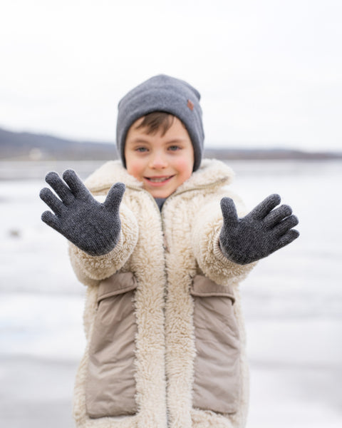 A young child with a happy expression, stands outdoors with both hands outstretched towards the viewer. They are wearing a grey knit beanie with a brown tag, a cream-colored sherpa coat with beige pockets, and dark grey knitted gloves. The background is a blurred, wintry landscape with what appears to be a frozen body of water.