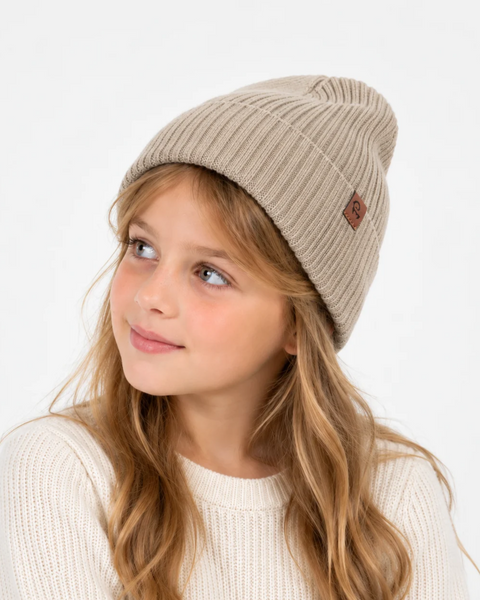 A young girl with long blonde hair wearing a ribbed beige knit beanie with a small brown patch logo, paired with a cream-colored sweater, against a plain white background.