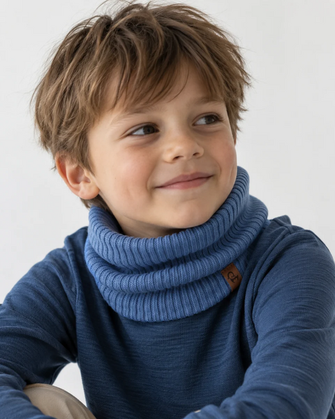 Young boy smiling while looking off-camera, wearing a ribbed blue knit neck gaiter and a matching blue long-sleeve top against a off-white background.