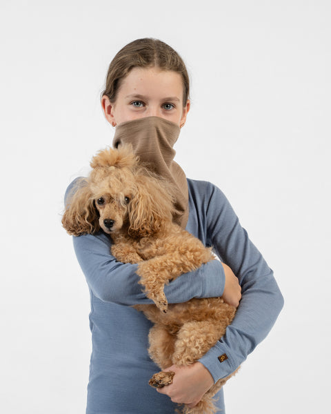 A young girl holding a small brown poodle dog in her arms while wearing a beige merino bandana pulled up over her nose and mouth.