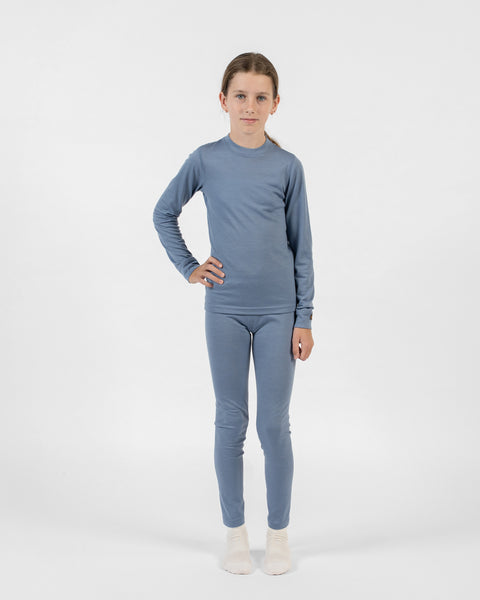 A full-body shot of a young girl facing forward, wearing a matching dusty blue merino long-sleeve top and bottoms set with cream socks, posing with her right hand on her hip.