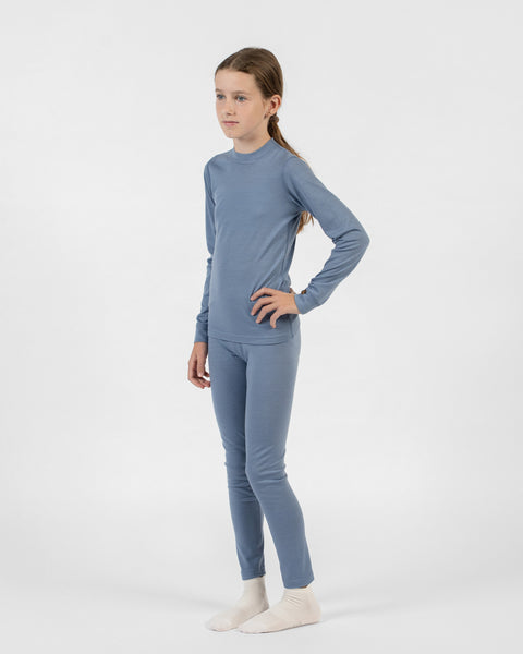 A full-body shot of a young girl turned slightly to the side, wearing a dusty blue merino long-sleeve and bottoms set, standing with her left hand on her hip and looking away from the camera.