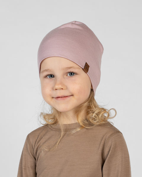 A studio portrait of a young blonde girl looking directly at the camera with a gentle smile. She is wearing a close-fitting dusty pink beanie hat featuring a small brown leather logo patch on the front edge. She is also wearing a light brown long-sleeved shirt. The background is a plain light grey.