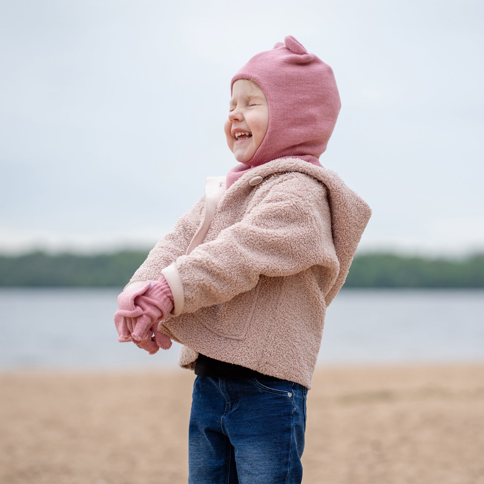 A young child with blue eyes and a wide, happy smile, is sitting cross-legged on a white surface. They are wearing a pink balaclava with ears, matching pink mittens, and a light-colored long-sleeved shirt and pants.