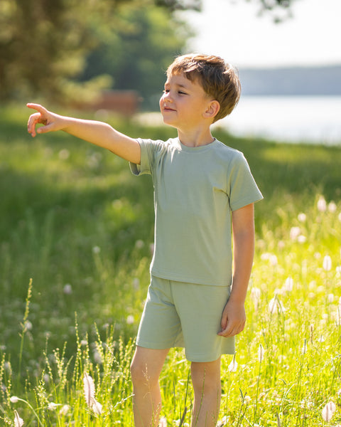 Young boy in a sunny meadow wearing a light green bamboo T-shirt and matching shorts. The lightweight kids' outfit is showcased in a natural outdoor setting, highlighting its soft, breathable comfort for everyday adventures.