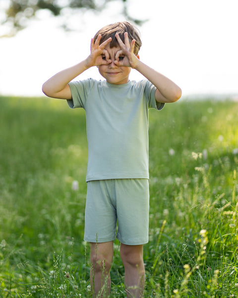Young boy standing in a sunny meadow wearing a light green bamboo T-shirt and matching shorts while playfully making binoculars with his hands. The lightweight kids' outfit is showcased in a natural outdoor setting, highlighting its soft, breathable comfort for everyday adventures.