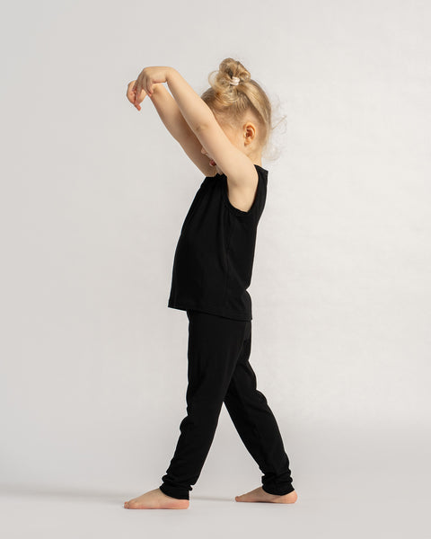 Child in a black sleeveless outfit standing sideways, stretching arms above head while barefoot against a light background.