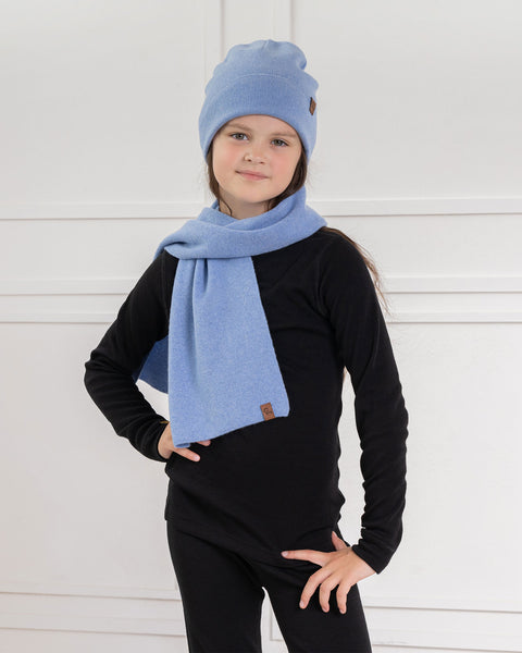 A child with long, dark hair, wearing a light blue knit beanie with a small brown tag and a matching light blue menique knit scarf, stands in front of a white wall with decorative molding. They are wearing a black long-sleeved shirt and black pants, with one hand on their hip and the other relaxed by their side. The child is looking directly at the camera with a neutral expression.