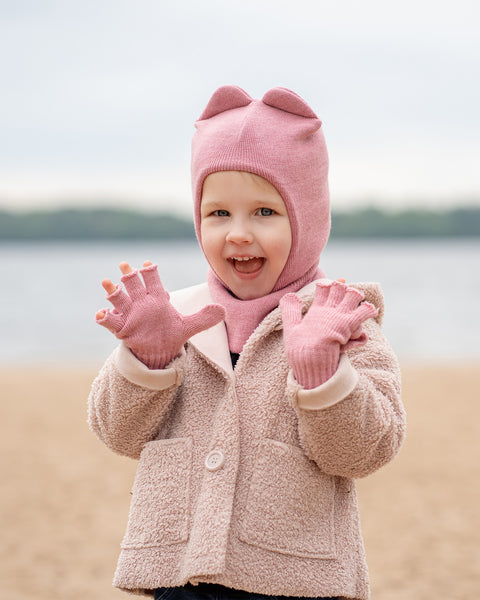 A joyful child wearing a pink balaclava with ears, a pink scarf, and a beige coat stands on a beach, hands open in excitement.