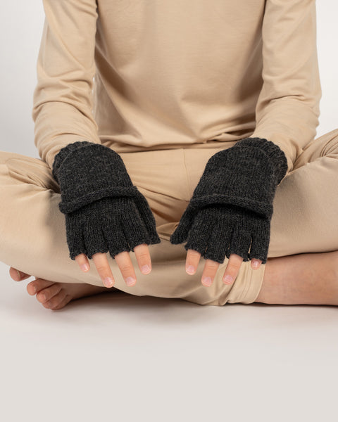Child sitting cross-legged wearing dark gray knitted convertible mittens, both mitten flaps open to reveal fingerless gloves.