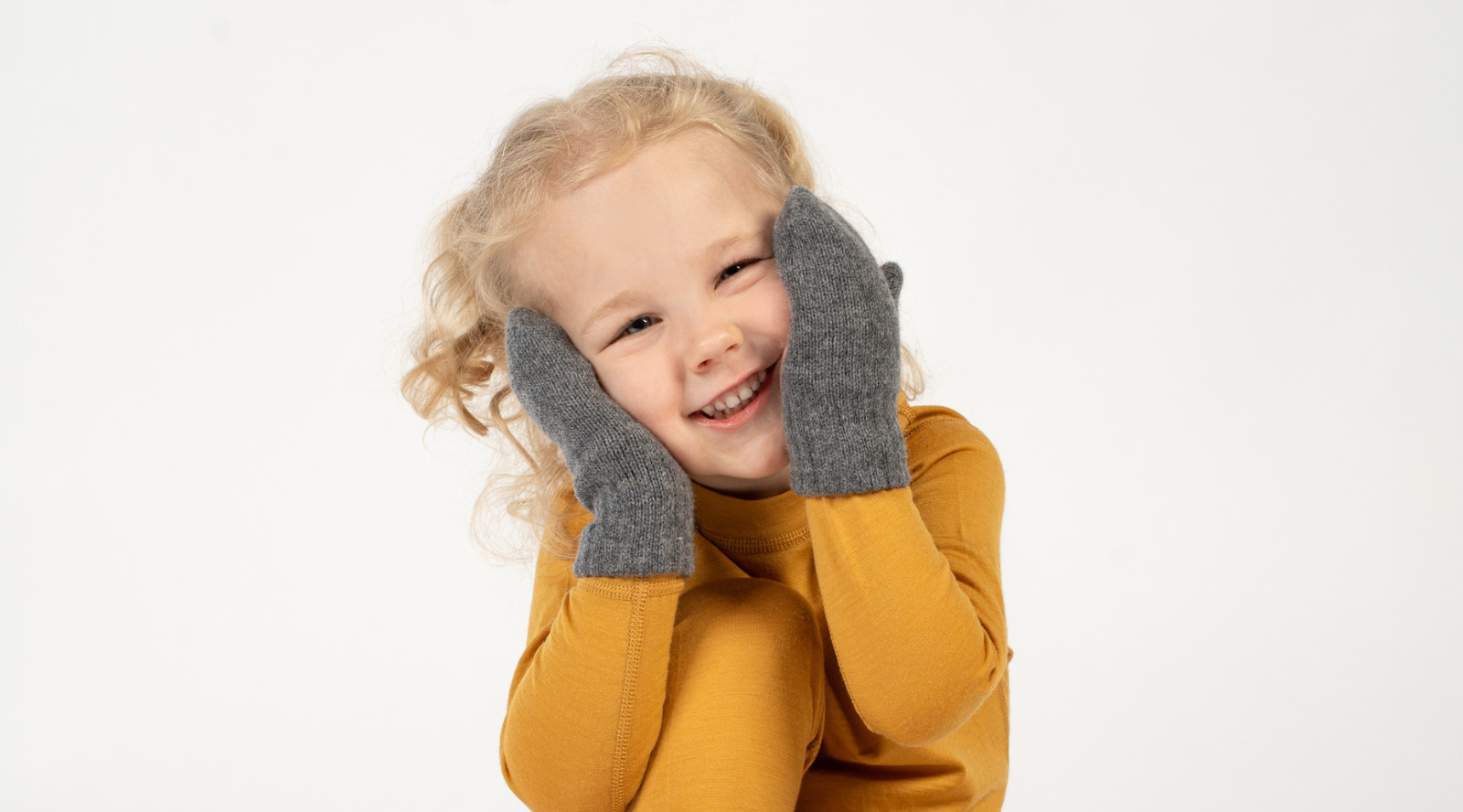A cheerful young child with blonde curly hair smiling brightly, wearing a mustard yellow long-sleeved top and framing their face with hands clad in grey knit mittens.