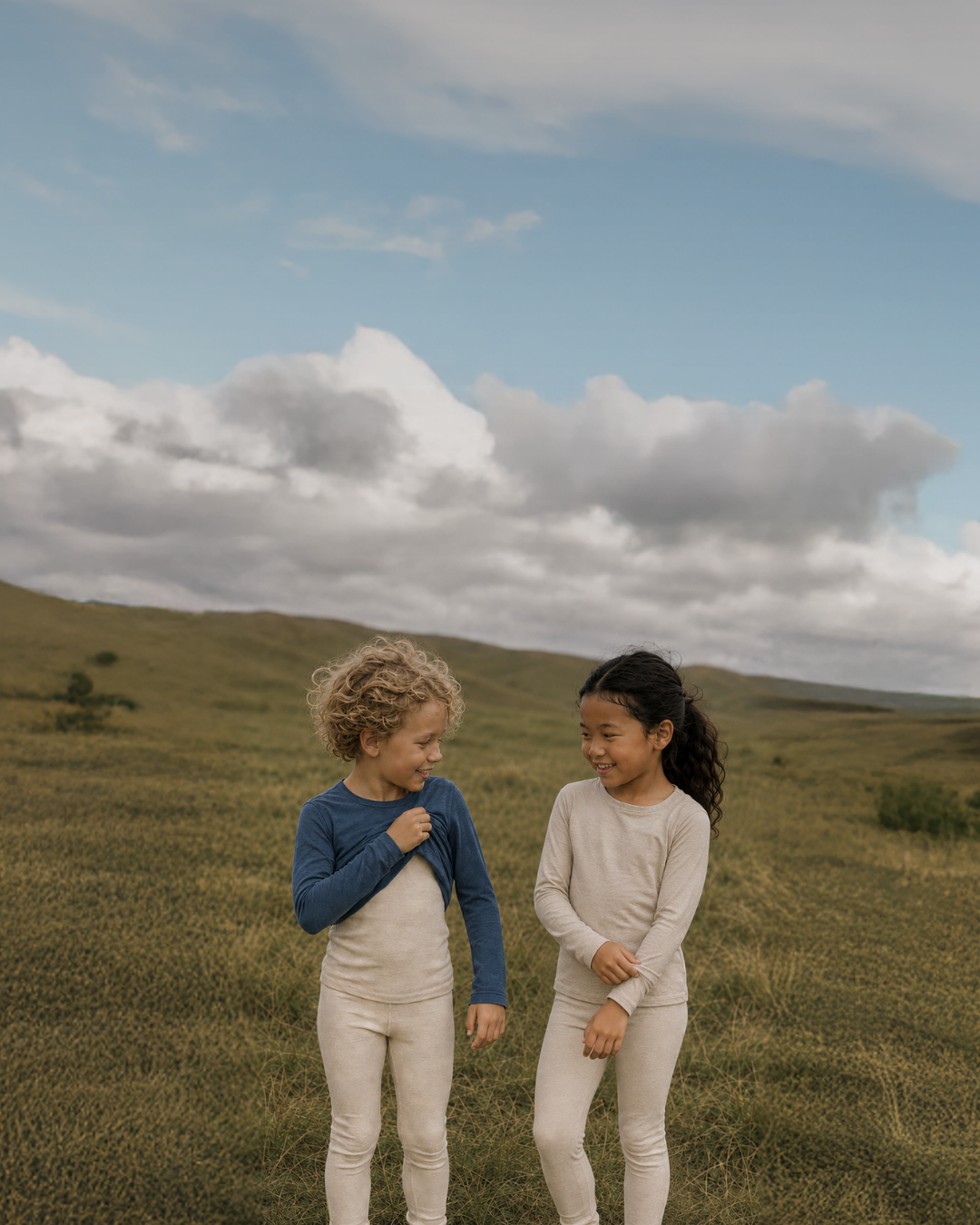 Two young children in long-sleeved base layers standing together in a grassy field under a partly cloudy sky.