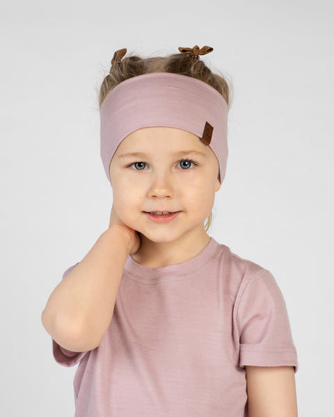 A close-up studio portrait of a smiling young girl looking directly at the camera with her hand gently resting on the back of her neck, against a white background. She is wearing a dusty pink short-sleeved t-shirt and a matching wide dusty pink headband featuring a small brown logo tag and ribbon tie details on top.
