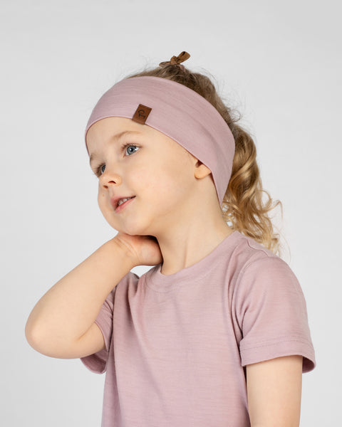 A close-up studio portrait of a gently smiling young girl looking off to her left, with her hand resting on the back of her neck against a white background. She is wearing a dusty pink short-sleeved t-shirt and a matching wide dusty pink headband with ribbon tie details visible at the top.