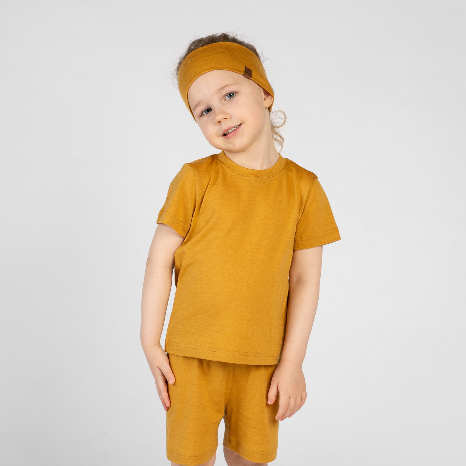 A close-up shot of a child's lower half standing barefoot in a grassy field with clover. The child is wearing simple mustard yellow shorts, with the hem of a matching yellow shirt visible at the top.