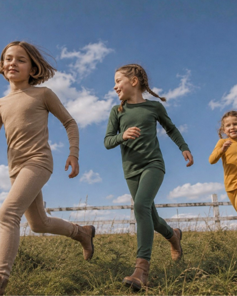 Three children running cheerfully through a grassy field under a blue sky with white clouds, wearing matching two-piece, long-sleeved thermal sets in beige, dark green, and mustard yellow.