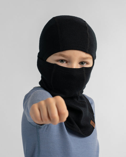 Young boy wearing a black knit balaclava face mask and blue t-shirt extending his fist toward the camera against a grey background.
