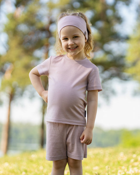 Young girl smiling outdoors wearing a dusty pink Merino wool T-shirt and a matching Merino wool headband. The soft, breathable accessories and clothing are showcased in a sunny natural setting, offering comfortable everyday wear for active ki