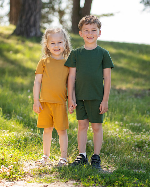 A young girl and boy stand smiling, holding hands in a sunlit grassy area near some trees. The girl, on the left, wears a mustard yellow t-shirt and shorts set with white sandals. The boy, on the right, wears a dark green t-shirt and shorts set with black sneakers.