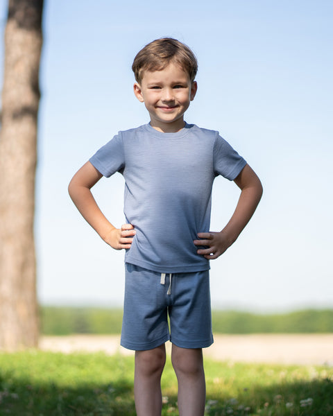 Young boy smiling outdoors wearing a dusty blue Merino wool T-shirt with a matching Merino wool shorts. The soft, breathable outfit is showcased in a sunny natural setting, offering comfortable everyday wear for active kids.