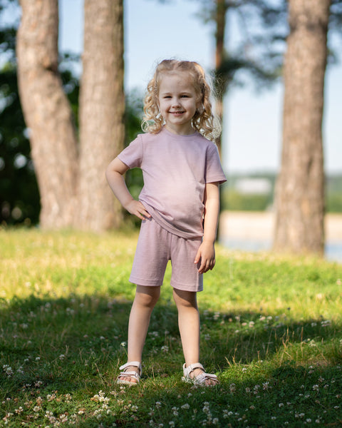 Young girl standing in a sunny park wearing a dusty pink Merino wool T-shirt and matching shorts. The lightweight, breathable kids' outfit is showcased in a natural outdoor setting, providing soft comfort for everyday play, travel, and warm-weather adventures.