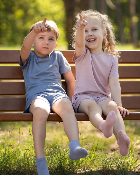 Two children sitting together on a wooden park bench wearing Merino wool T-shirts and matching shorts in dusty blue and dusty pink. They point excitedly into the distance, showcasing the lightweight, breathable kids' outfits in a sunny outdoor setting perfect for everyday adventures.