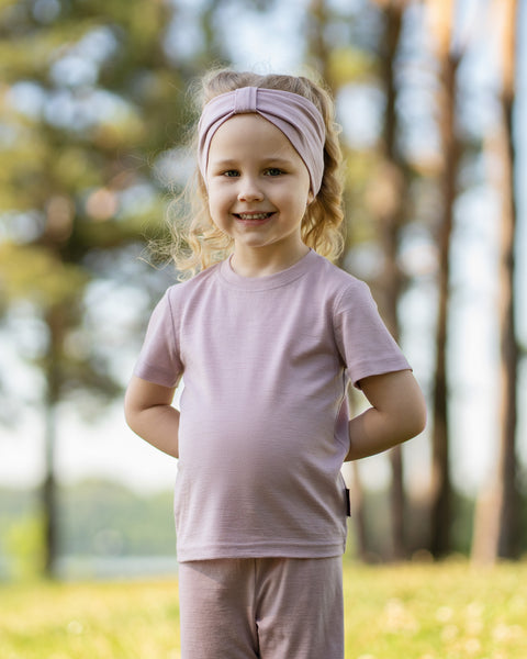 Young girl smiling outdoors wearing a dusty pink Merino wool T-shirt and a matching Merino wool headband. The soft, breathable accessories and clothing are showcased in a sunny natural setting, offering comfortable everyday wear for active ki