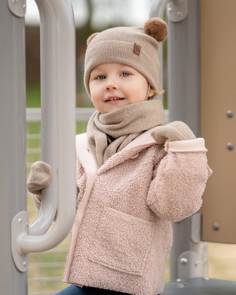 A young child, dressed in a pink textured jacket with a hood, a creamy beige menique knit beanie with two brown pom-poms on top and a small brown tag, a matching scarf, and meinque mittens, stands on a playground structure. The child is looking directly at the camera with a slight smile. Their hands are gripping a light gray metal bar. The background is blurred with hints of green and brown.