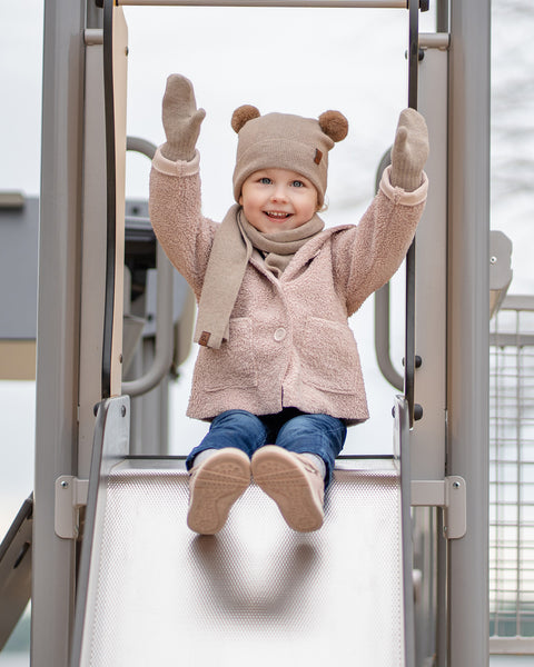 A young child with blue eyes and a joyful expression, is seated at the top of a metal playground slide. They are wearing a beige balaclava with brown pom-pom ears, a matching beige scarf, beige mittens, a beige sherpa-style coat, blue jeans, and pink sneakers, with their arms raised in excitement.