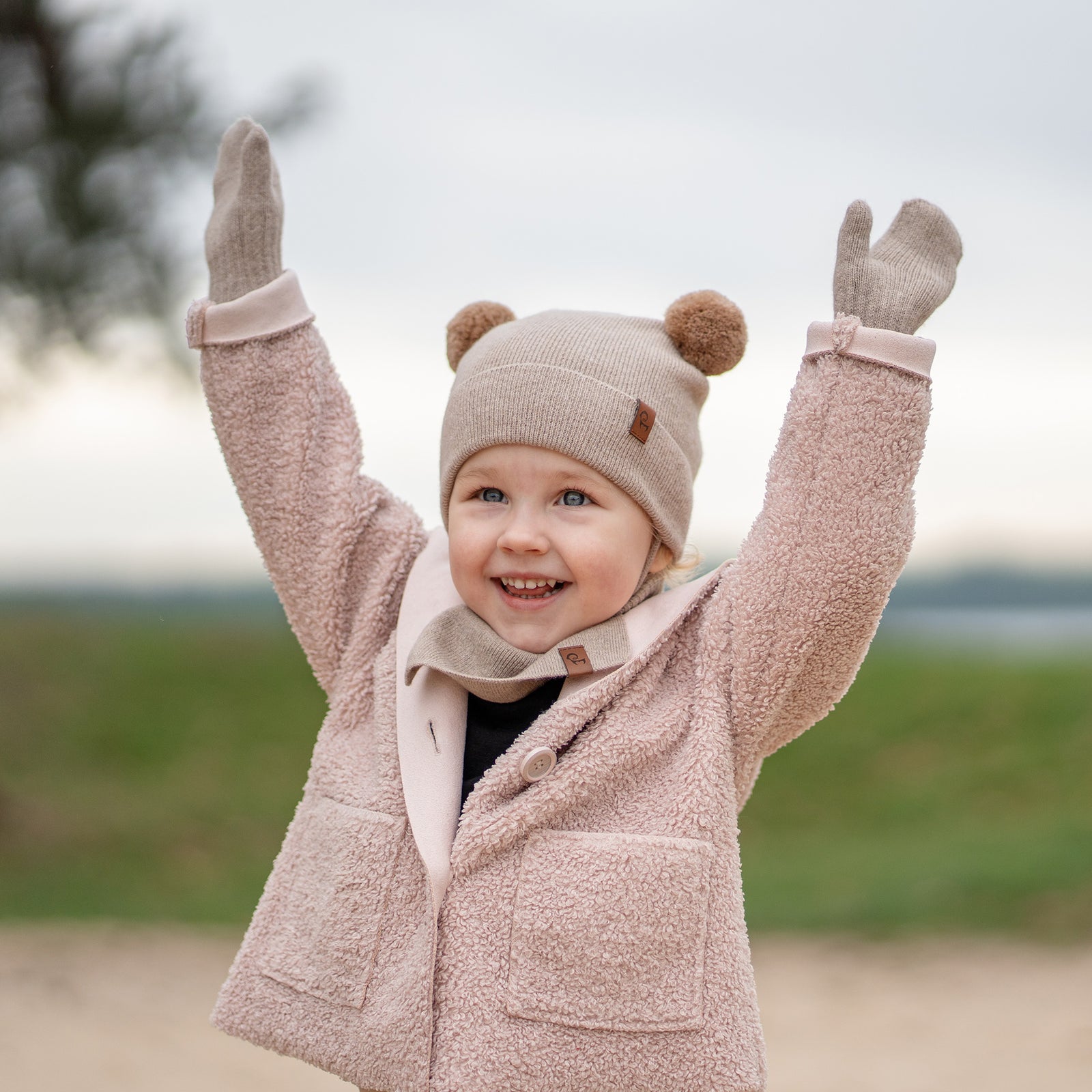 A young child with light brown curly hair and a subtle smile, is looking directly at the viewer. They are wearing a light blue knit hat with two pom-poms and earflaps that tie under the chin, matching light blue mittens, and a light blue knit neck warmer, over a black long-sleeved shirt. The background is a white paneled wall.