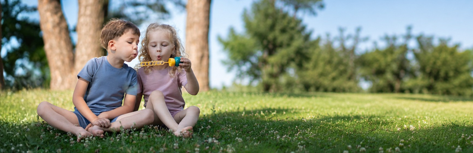 Two children sitting on a grassy lawn in a sunny park, playing with a small toy airplane. The boy wears a blue t-shirt and shorts, and the girl wears a pink t-shirt and shorts.