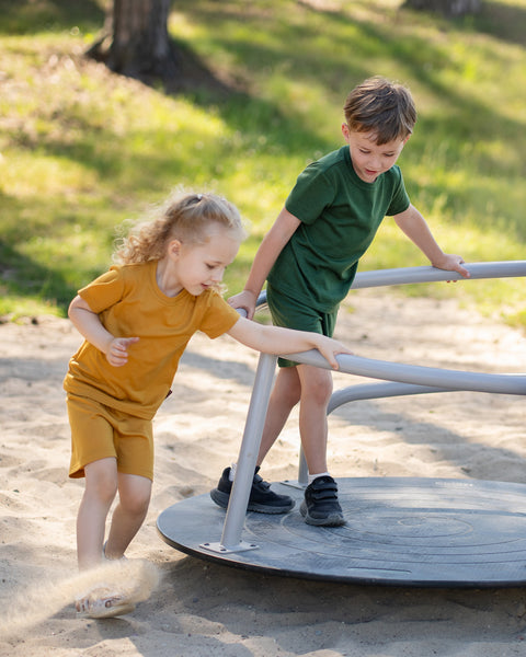 Two children playing together on a playground merry-go-round while wearing matching Merino wool T-shirts and shorts in yellow and dark green. The lightweight, breathable kids' outfits are showcased in a sunny outdoor setting, perfect for active play, everyday comfort, and family adventures.