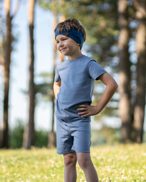 Young boy smiling outdoors wearing a dusty blue Merino wool T-shirt with a matching Merino wool shorts and headband. The soft, breathable outfit and accessory are showcased in a sunny natural setting, offering comfortable everyday wear for active kids.