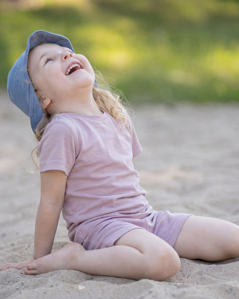 Young girl sitting on a sandy beach wearing a dusty pink Merino wool T-shirt, matching shorts, and a dusty blue sun cap hat. The lightweight, breathable kids' outfit is showcased in a sunny outdoor setting, making it ideal for summer adventures, travel, and everyday comfort.