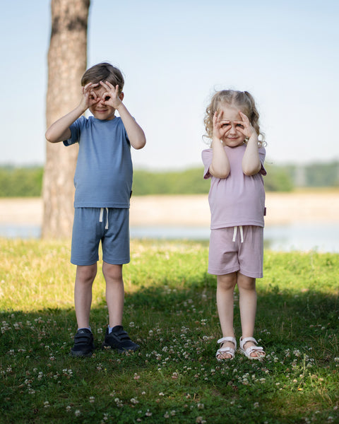 Two children standing together on the grass wearing Merino wool T-shirts and shorts while blowing bubbles on a sunny day. The lightweight, breathable kids' outfits are showcased in a playful outdoor setting, perfect for everyday comfort and active adventures.