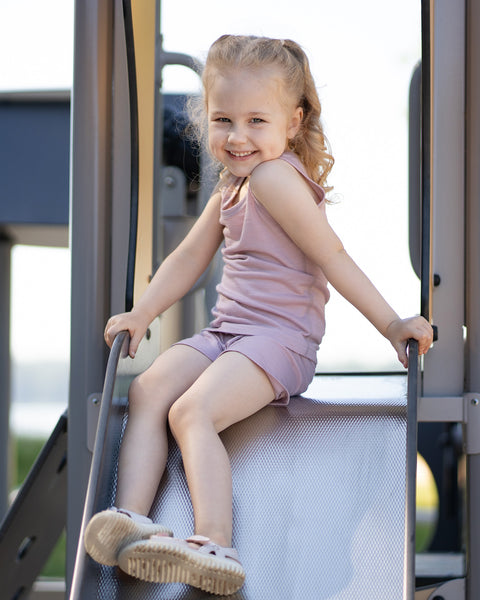 A smiling young girl with blonde hair tied half-up sits on a metal playground slide, looking back over her shoulder at the camera. She is wearing a matching dusty pink tank top and shorts set.