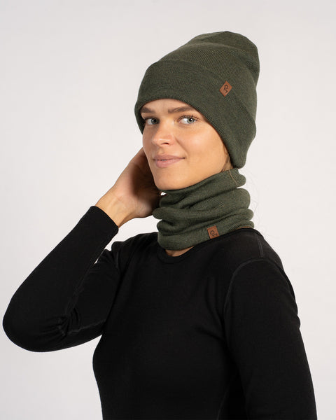 Woman wearing a dark green knit beanie and a matching neck gaiter, dressed in a black long-sleeve top, smiling softly with one hand touching her hair against a plain light background.