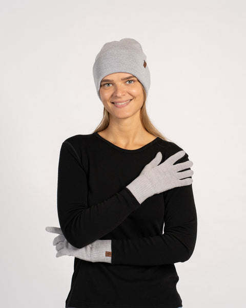 Woman wearing a light gray knit beanie and matching gloves, dressed in a black long-sleeve top, smiling with arms crossed and one gloved hand resting on her shoulder against a plain background.