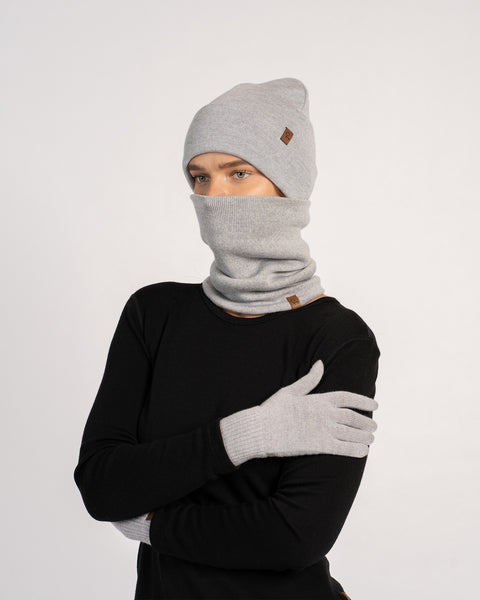 Woman wearing a light gray knit beanie, matching gloves, and a neck gaiter covering her face, dressed in a black long-sleeve top, posing with arms crossed against a plain background.