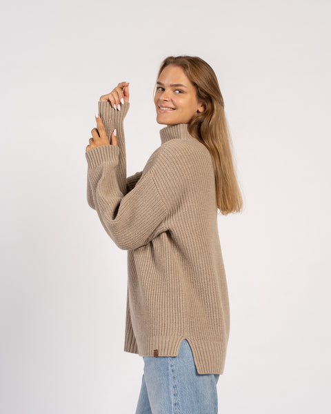 A woman smiling while wearing a loose beige knit turtleneck sweater with side slits, paired with light blue jeans.